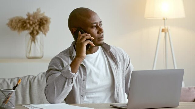 A millennial African American man in his 30s is engaged in a phone conversation while seated at his home office. He is focused, with a laptop open in front of him and papers scattered on the table.
