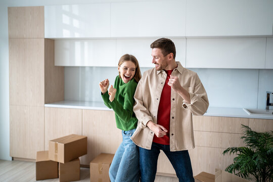 Loving family couple have fun on moving day. Happy, overjoyed man and woman smiling dancing in empty kitchen with cardboard boxes in new purchased apartment. Pleasure of fulfilling dream goal together