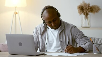 A man in his 30s focuses intently on his laptop during a virtual session from his living room. He takes notes and listens attentively while wearing a headset, showcasing a modern lifestyle.