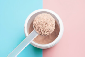 Chocolate protein powder in scoop against plastic jar on pink and blue background.