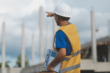 Asian engineer handsome man or architect looking forward holding paperwork blueprint with white safety helmet in construction site. Standing at modern building.