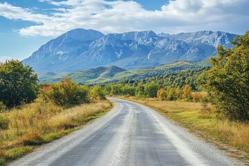 Fototapeta premium Scenic asphalt road winding through autumnal landscape towards majestic mountains under a partly cloudy sky.