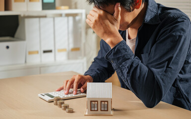 Stressed person holding a calculator, with stacks of coins and a house model nearby. Reflects...