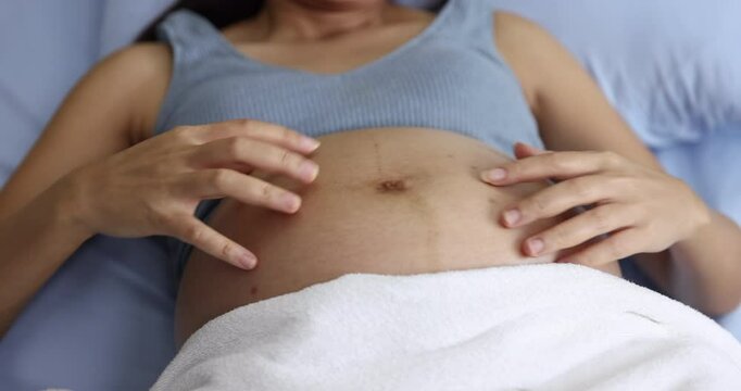 A pregnant woman gently scratching or soothing an itch on her baby bump while resting on a bed, highlighting the physical changes and self-care routines associated with pregnancy