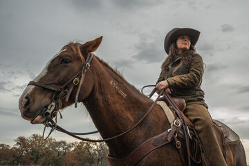 Obraz premium Woman Wearing Cowboy Hat and Looking Off While Sitting On Brown Horse With Cloudy Sky