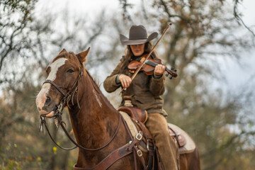 Fototapeta premium Woman Looking Down Wearing Cowboy Hat and Playing Fiddle Violin While Sitting On Brown and White Horse In Front of Trees