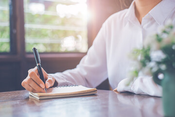 Close-up a woman's hand writing on a notepad with a black pen on a wooden desk in a window-side office.