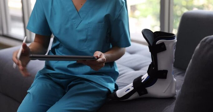 A medical professional wearing scrubs sits on a couch holding a tablet, with a white orthopedic boot placed beside them, symbolizing healthcare, recovery, and digital patient management in a relaxed s