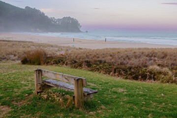 Empty beach at dawn, volleyball net on the sand, wooden bench on grassy hill. Tranquil coastal scene. ONEMANA, COROMANDEL PENINSULA, NEW ZEALAND
