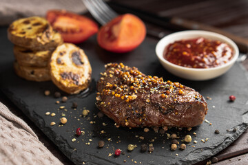 Roasted pork steak on black stone board over dark background. With tomato, sauce and fried potato