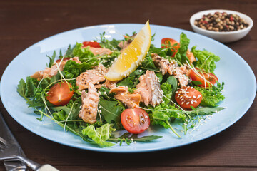 Salmon salad with green leaves, tomato, lemon, sesame in blue plate on wooden table