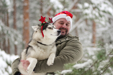 positive Caucasian man in a Santa hat and his husky dog with a red decoration on his head celebrate Christmas.
