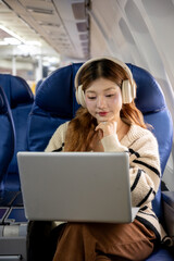 Asian woman sitting watching a movie on her laptop on an airplane while traveling somewhere during her vacation.