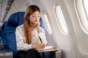Fototapeta premium A focused Asian businesswoman works during the flight, taking notes in a book, flying abroad.
