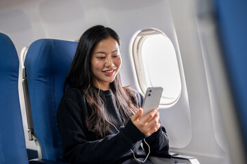 A relaxed, happy Asian woman using her smartphone and listening to music during the flight.