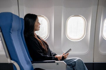 Relaxed Asian female passenger sits at a window seat, looking at the view outside during the flight