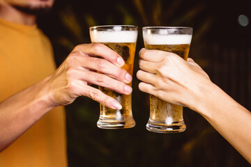 A man's and woman's hands raise beer glasses group in a lively night party, surrounded by family and friends. The evening is filled with warmth, laughter, and joyful celebration.