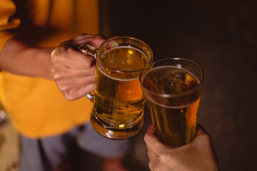 A man's and woman's hands raise beer glasses group in a lively night party, surrounded by family and friends. The evening is filled with warmth, laughter, and joyful celebration.