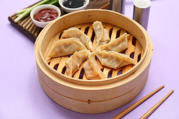 Bamboo steamer with tasty Japanese gyoza and bowls of sauces on lilac background