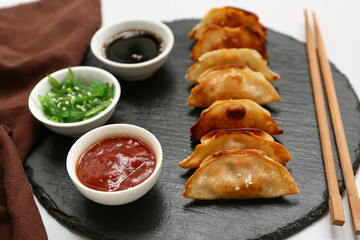 Black board of fried Japanese gyoza with bowls of sauces and seaweed salad on grey background