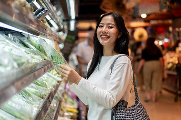 A smiling Asian woman grocery shopping at a supermarket, selecting fresh vegetables from a shelf.