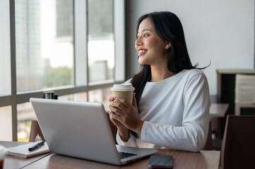 An Asian woman enjoying a hot drink at a coffee shop while taking a break from work on her laptop.