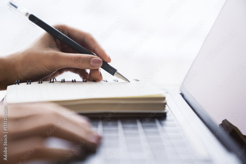 Poster in close view, a woman's hand writing down notes in a notepad on a laptop, against a hazy backdrop