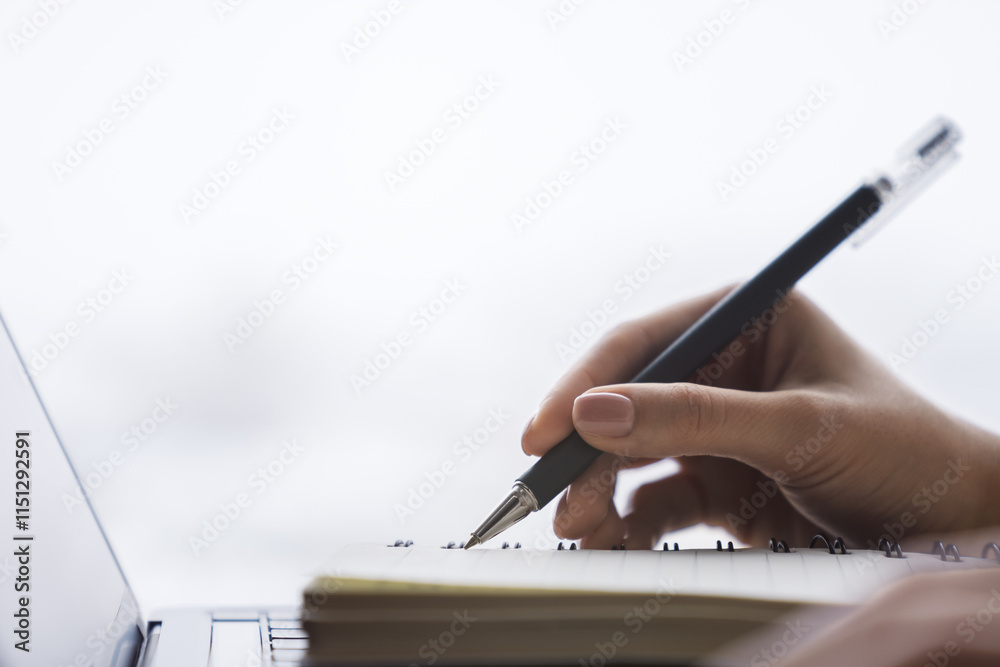 Poster in close view, a woman's hand writing down notes in a notepad on a laptop, against a hazy backdrop