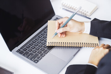 A woman's hand seen in close-up, writing in a notepad on a contemporary laptop, against a soft-focus backdrop