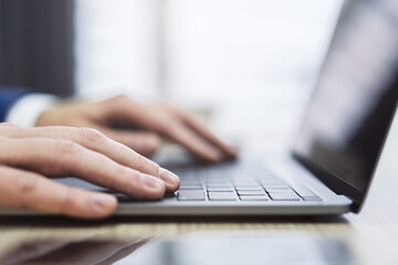 With an office scene appearing softly blurred in the backdrop, the picture shows a close-up of male hands typing on a contemporary laptop keyboard