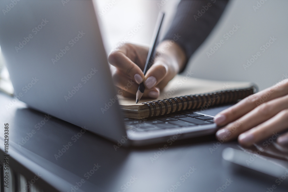 Poster close perspective of a woman's hand scribbling in a notepad on a stylish laptop, with a soft blur in