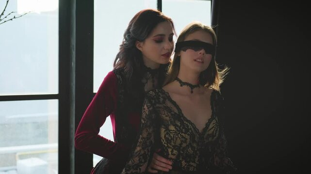 Two beautiful women. Evil angry vampire queen is tempting enchant embrace, hug young scared princess. Black blindfold on eyes. Long hair fluttering fly wind. Red black halloween costume. Horror woman