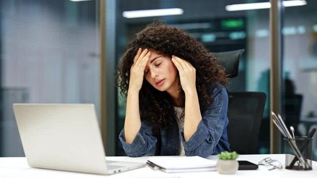 Tired businesswoman with curly hair sitting at desk in office rubbing temples. Exhausted, overworked female suffering from headache, chronic migraine in workplace.