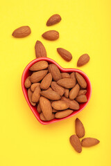 Fresh natural healthy almonds nuts in heart shaped bowl on yellow background. Vertical photo.