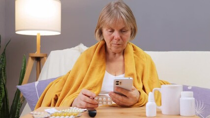 An elderly woman reviews her medication schedule using her phone while wrapped in a warm blanket at home. She appears focused, surrounded by various health items on a table