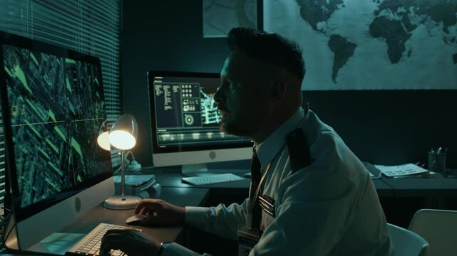 Medium side shot of male military officer in uniform working night shift alone at desk, monitoring satellite surveillance, detecting threat, zooming in and peering at computer screen with concern