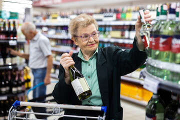 Mature woman with glasses chooses bottle of wine in alcohol section of supermarket