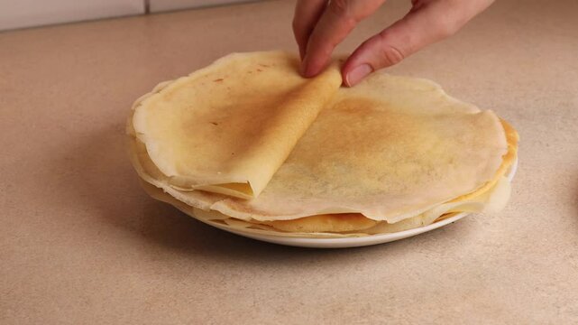 Woman peeling thin crepe from stack on plate. Soft, golden pancakes made fresh for breakfast or dessert. Perfect for sweet or savory fillings, showcasing homemade cooking.