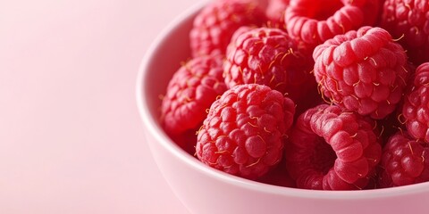 Close up view of a bowl filled with ripe fresh raspberries against a soft pink background, highlighting the vibrant red color and juicy texture of these delicious raspberries.