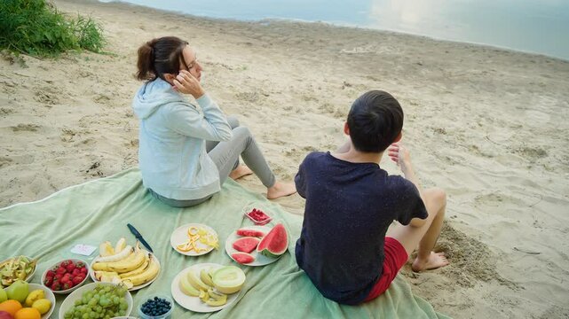 Mother and son eating raspberry at healthy food picnic near calm riverside. Woman savoring fresh berry while sitting on green blanket next to boy. Lady and young male surrounded by colorful fruit