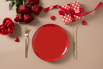 Valentine's day. Romantic dinner for date with red plate, chocolate sweets, romantic gift and red roses on beige background. View from above. Copy space.
