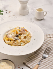 plate of fettuccine topped with creamy sauce and sprinkled with black pepper and herbs is served on a white table. Small container holds extra sauce nearby.