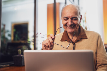 Cheerful senior man in casual clothing holding eyeglasses in his hand and smiling while using laptop in the cafe.