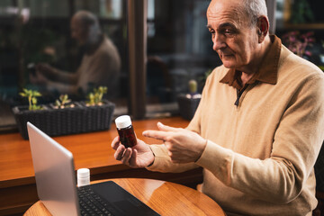 Senior man showing bottle of pills and talking with his doctor during a video call on laptop while sitting in the city cafe.