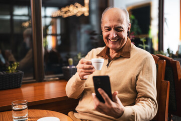 Happy senior man drinking coffee while using mobile phone and earphones. He is relaxing in the cozy city bar at evening and using technology.