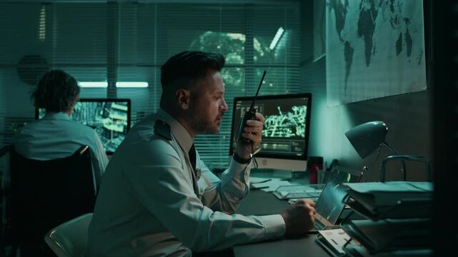 Medium side shot of Caucasian male military satellite reconnaissance officer working at night in operations centre, monitoring satellite footage on laptop, talking to colleagues on portable radio