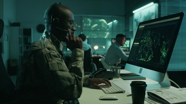 Medium side shot of African American male military satellite operator in camouflage uniform working at night in command center, monitoring surveillance footage, answering phone call on headset
