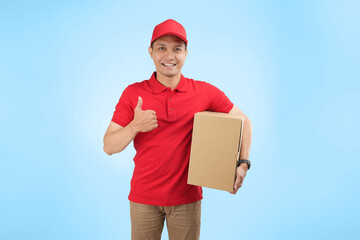 An happy Asian courier gift a thumb while holding a package against a blue background wearing red company uniform.