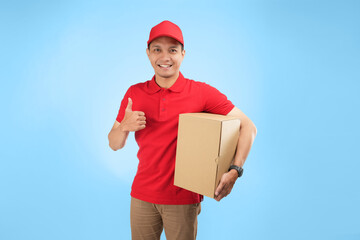An happy Asian courier gift a thumb while holding a package against a blue background wearing red company uniform.