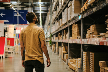 Young man stands in warehouse aisle, looking at rows of shelves stocked with cardboard boxes. Rear view. Warehouse well-lit, spacious. Modern distribution center.